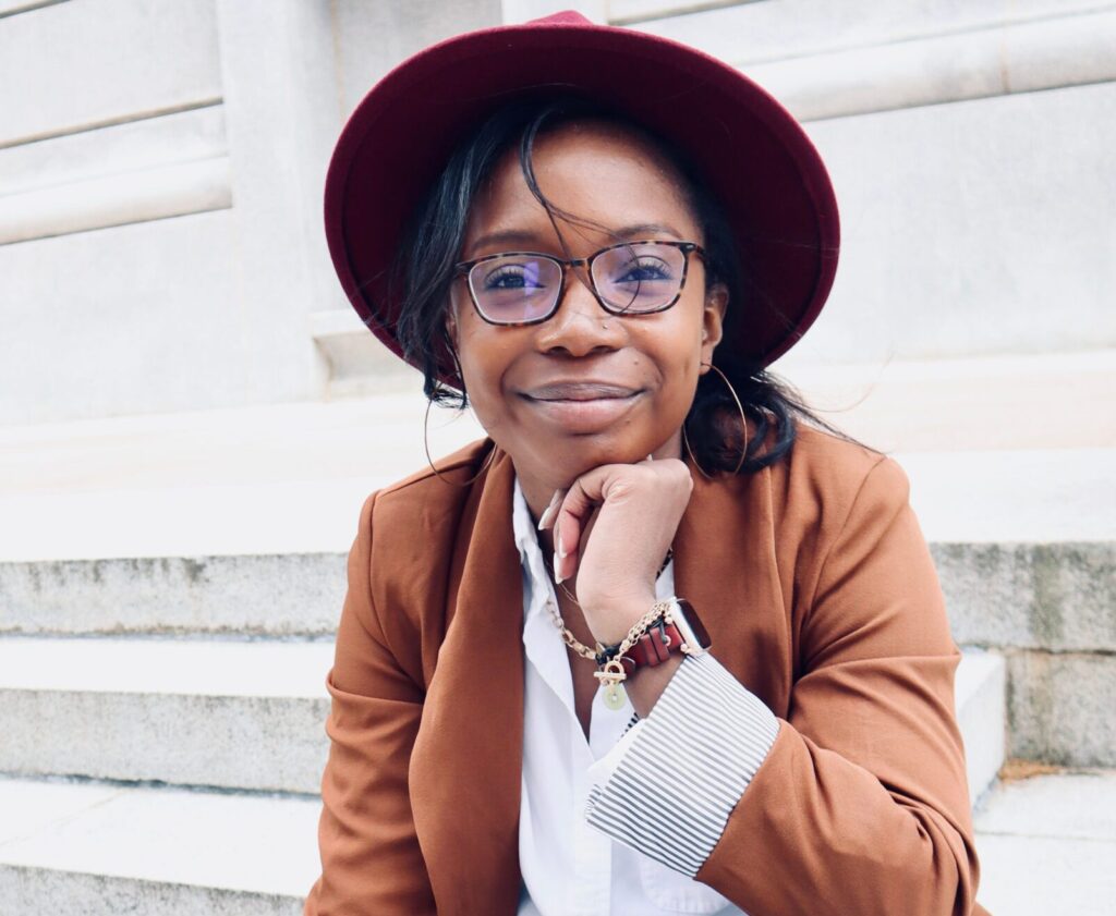 Erica, who is black is wearing glasses with her hand under her face, wearing a white blouse and brown blazer and marood hat