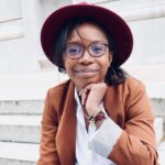 Erica, who is black is wearing glasses with her hand under her face, wearing a white blouse and brown blazer and marood hat