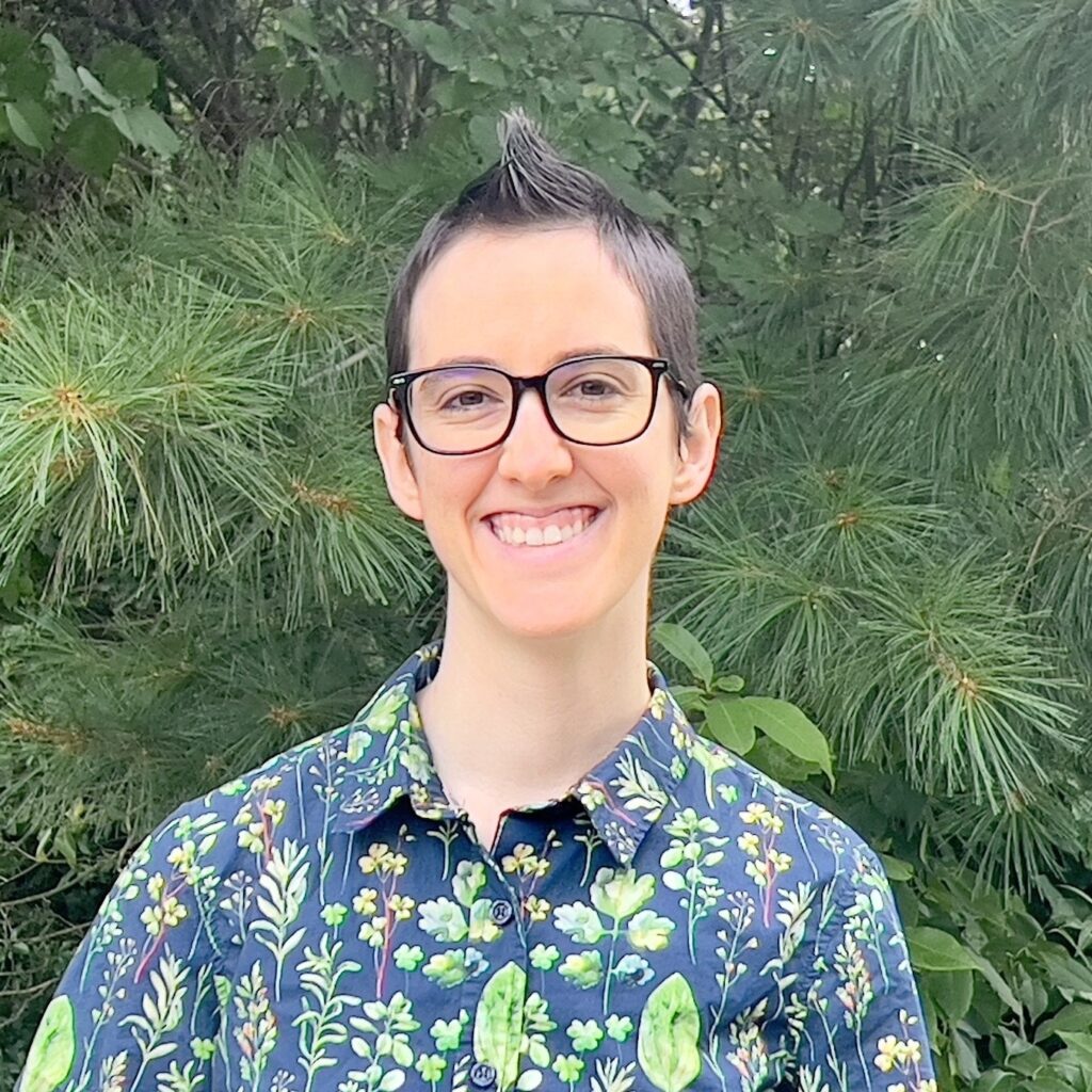 Claire MR photo square A pale genderfluid person with a fauxhawk, wearing a blue shirt with green watercolor plants on it. The background is pine trees.