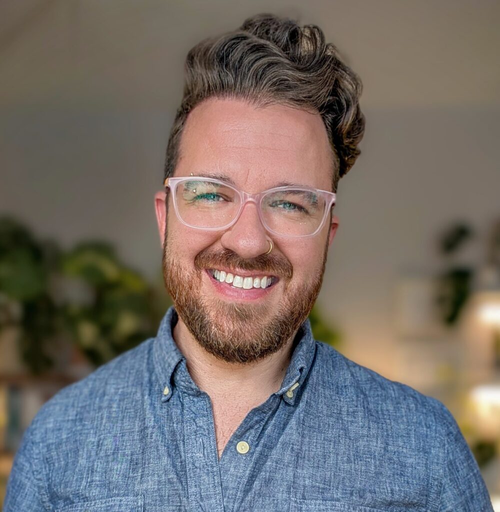 2025-08 Michael Kuffel Portrait A head-and-shoulders portrait of an AMAB nonbinary person with a beard, glasses, and a nose ring. They have brown, wavy hair styled to the side and is wearing a blue collared shirt. They are smiling warmly at the camera.