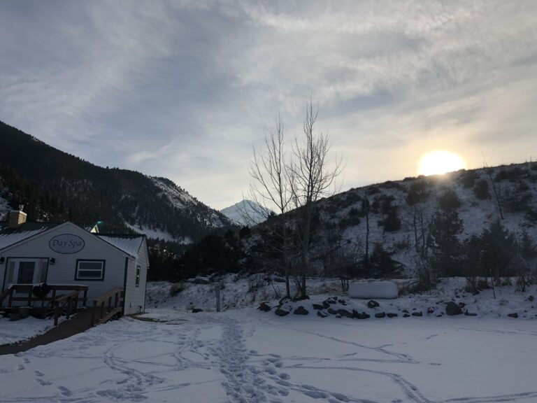 Snow-covered mountain landscape at sunrise with a small white building in the foreground, and footprints crossing a snowy open area. Pine-covered hills and bare trees surround the building, with soft winter light illuminating the scene.