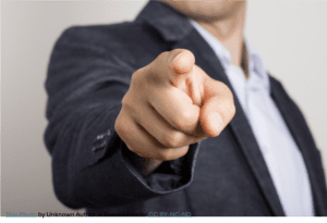 Close-up of a person in a dark suit jacket pointing directly at the camera, with the finger in sharp focus and the face out of the frame and blurred in the background.