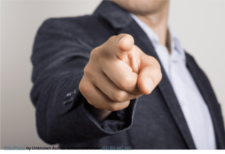 Close-up of a person in a dark suit jacket pointing directly at the camera, with the finger in sharp focus and the face out of the frame and blurred in the background.
