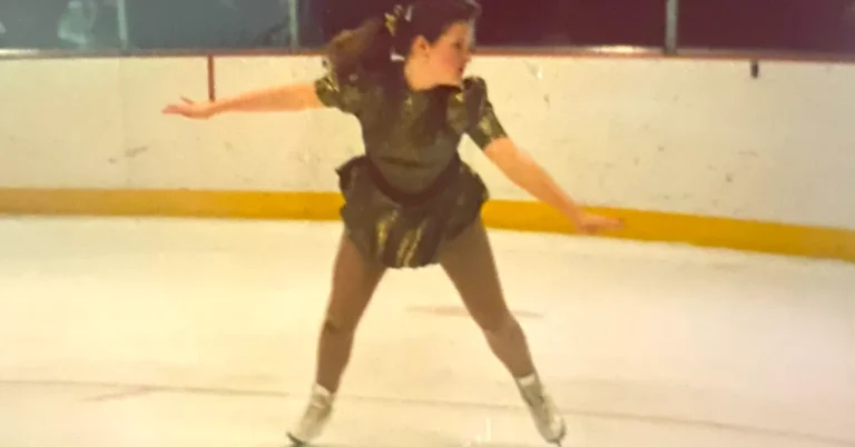 A figure skater wearing a dark green dress glides across an indoor ice rink with her arms extended to the sides. She appears mid-movement, looking to her left, with white skates on the ice and rink boards visible in the background.