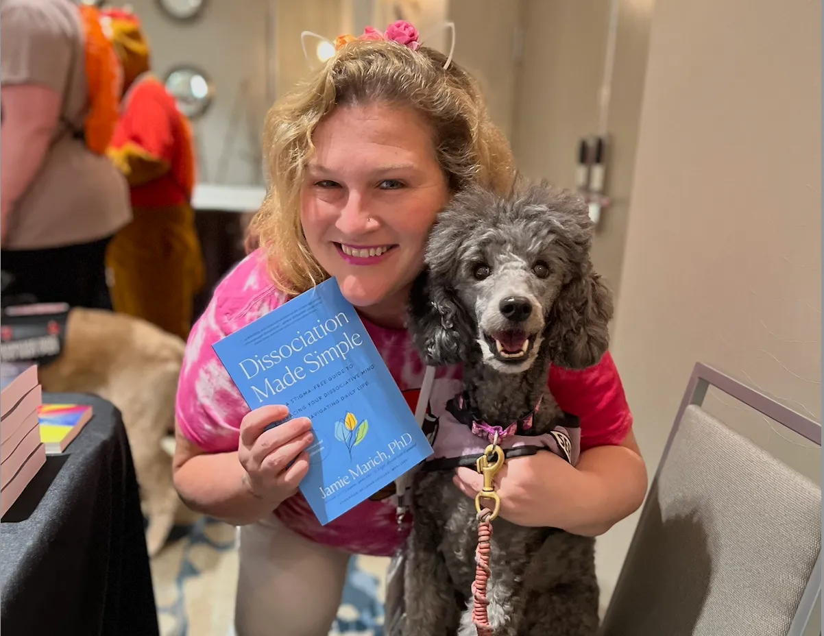Jamie Marich smiles while wearing a pink tie-dye tshirt and headband with pink flowers and cat ears. Holding in her right hand her book Dissociation Made Simple, in her left arm is a gray poodle, Starlight, who is wearing a pink harness and leash who is looking at the camera.