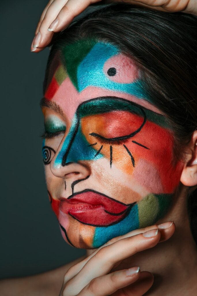 Close-up portrait of a person with eyes closed, their face painted in a vibrant abstract design resembling cubist art. Bold blocks of teal, red, orange, pink, blue, and green cover the face, outlined with black lines that create stylized features, including an exaggerated eye shape around one eyelid and a spiral motif on the cheek. One hand rests on the forehead while another supports the chin against a dark, blurred background.