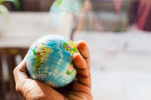 A hand holds a small squeeze ball shaped like a globe. The globe shows South America, the Atlantic Ocean, and part of Africa. The background is blurry and looks like an outdoor space.