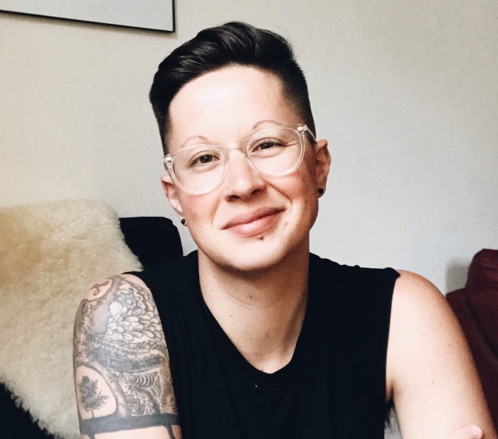 Korrine is sitting in a black chair with a cream colored fuzzy blanket hanging on the back of it. They are wearing a black sleeveless shirt and clear glasses. They have short brown hair and are gently smiling at the camera.