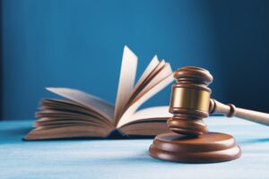 An open book with pages fanning out sits in the background, while a wooden judge's gavel rests in the foreground on a light blue surface against a dark blue backdrop.