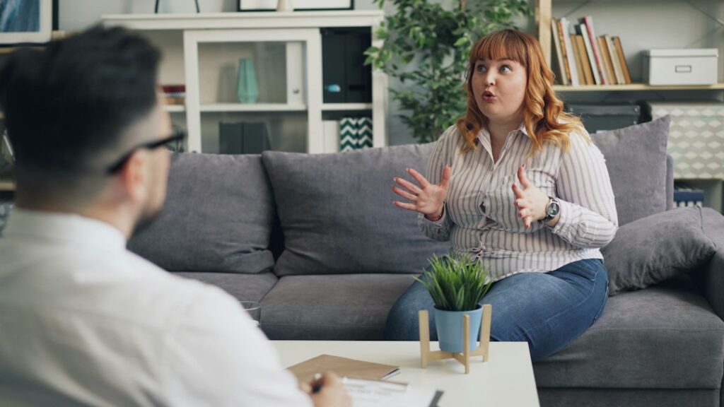 A woman sits on a sofa gesturing with her hands while talking to a person sitting opposite her in an office setting.