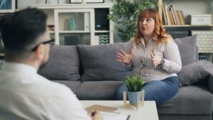 A woman sits on a sofa gesturing with her hands while talking to a person sitting opposite her in an office setting.